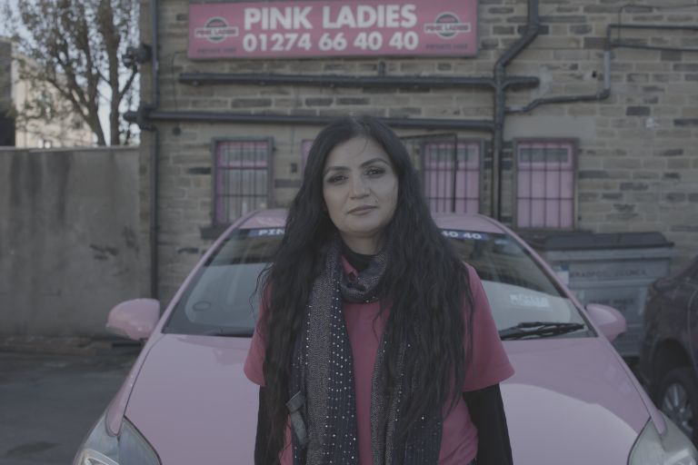 A medium shot of a South Asian woman. She is wearing a long, softly draped scarf styled around her shoulders, looking directly toward the camera with a warm, confident expression. She is stood infront of a pink stationary car, and on the building behind her is a sign that says Pink Ladies.