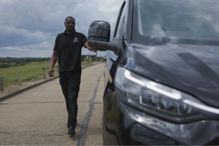 A taxi driver wearing all black walks towards his black shiny taxi. In the background there is a road and a green bit of parkland.