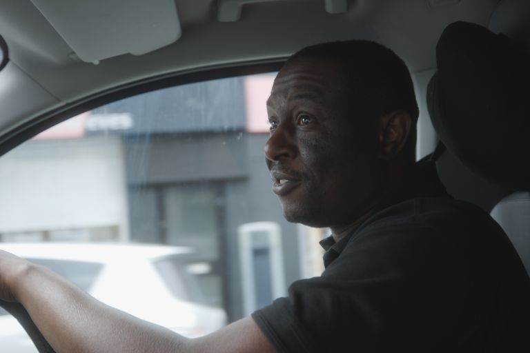 A black taxi driver sat in the drivers seat inside his taxi.