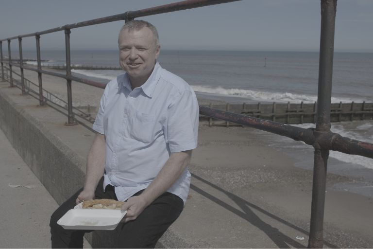 A taxi driver wearing a light blue shirt and black trousers is sat on a wall infront of black railings. On his lap is fish and chips in a white carton. The sea is grey behind him.